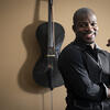 Subject posing and smiling in front of his cello, which is hanging on a wall behind him